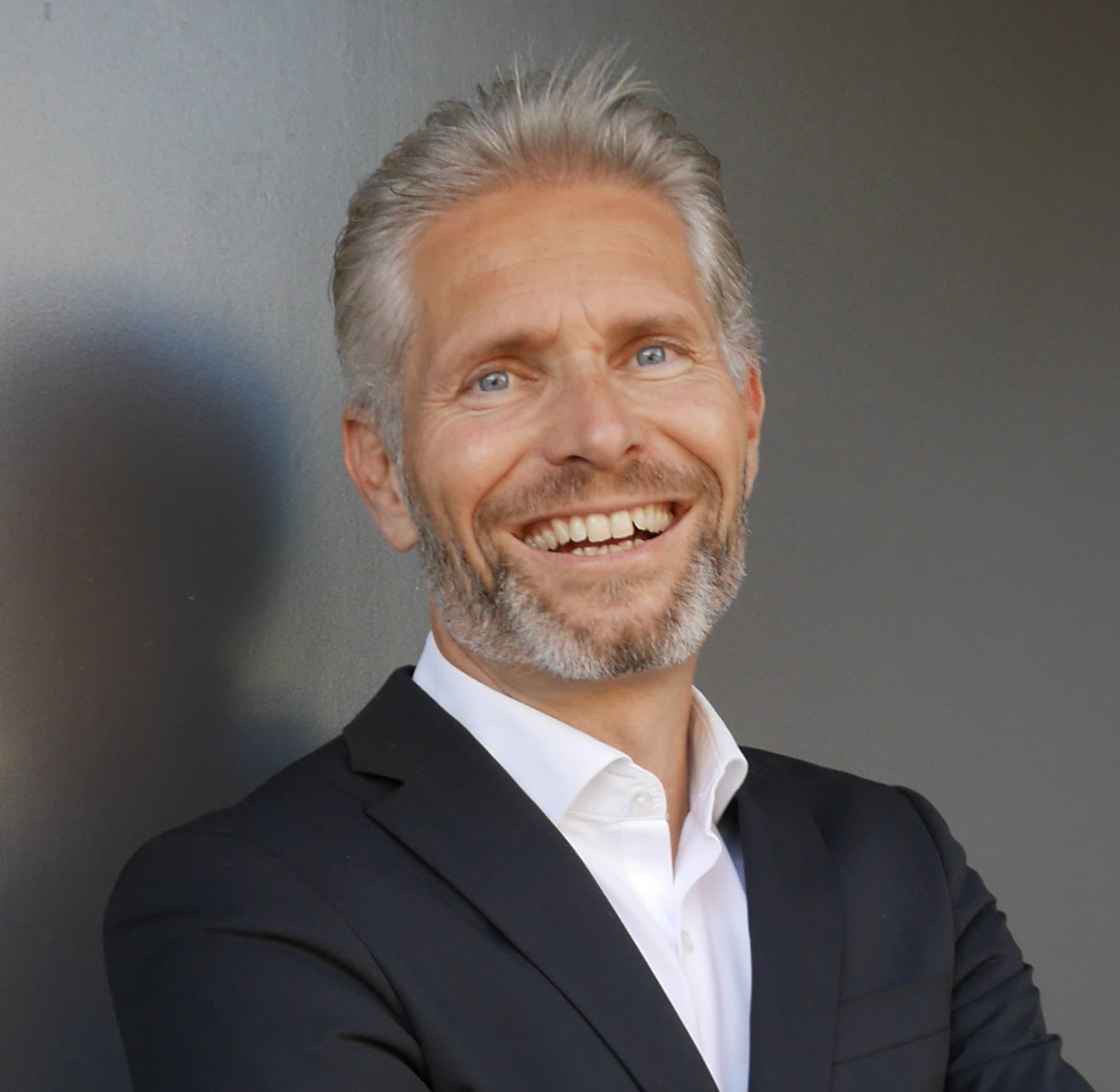 Claude Honegger, a mid-aged male with grey hair and beard, smiling in front of a grey background.
